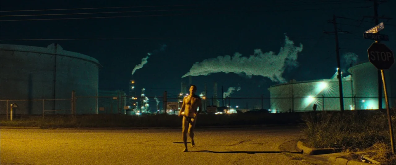 Red Rocket (2021) movie still: wide — A nighttime industrial area features tall storage tanks and smoking chimneys behind a fence. A shirtless man…