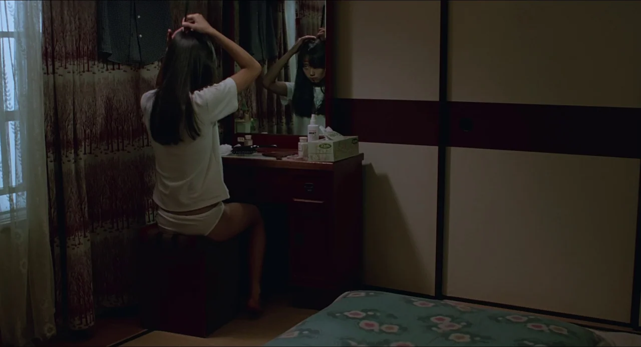 Typhoon Club (1985) movie still: medium — A dark, modest room features a wooden vanity with a mirror and sliding doors. A young woman sits on a…