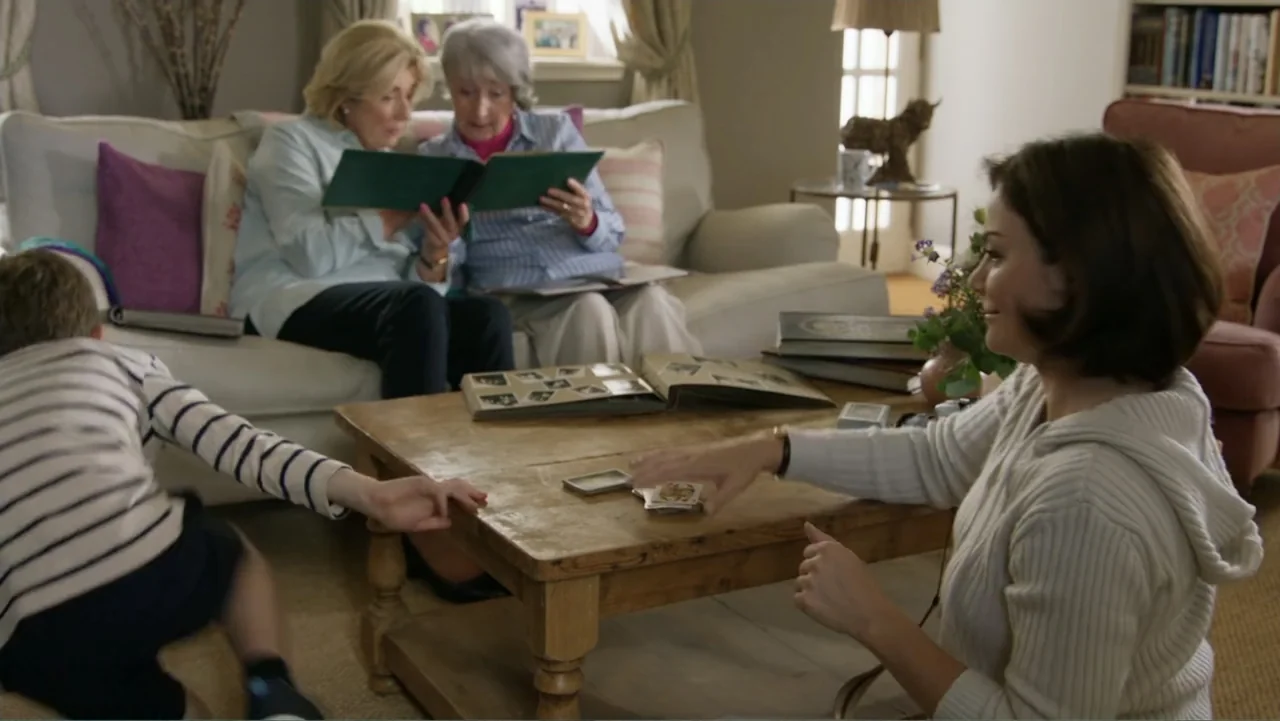 A Scottish Love Scheme (2024) movie still: medium — A well-lit domestic space contains a seating area with a sofa and coffee table. A woman and…