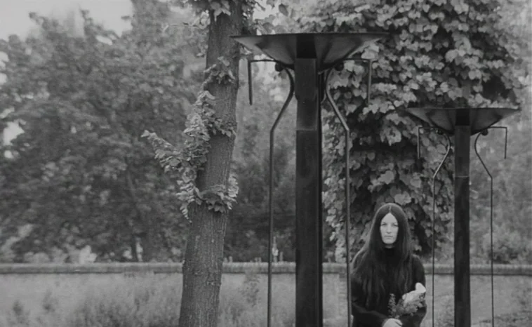 The Cremator (1969) movie still: medium — A woman stands between two metallic sculptural pillars in a dense outdoor park area. She faces forward…