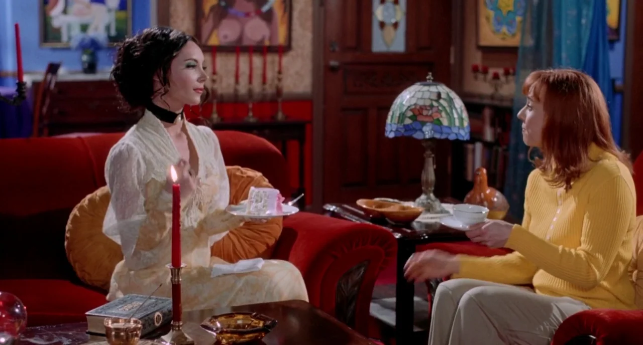 The Love Witch (2016) movie still: medium — Two women interact in a colorfully decorated living room with red upholstery. One woman holds a plate of…