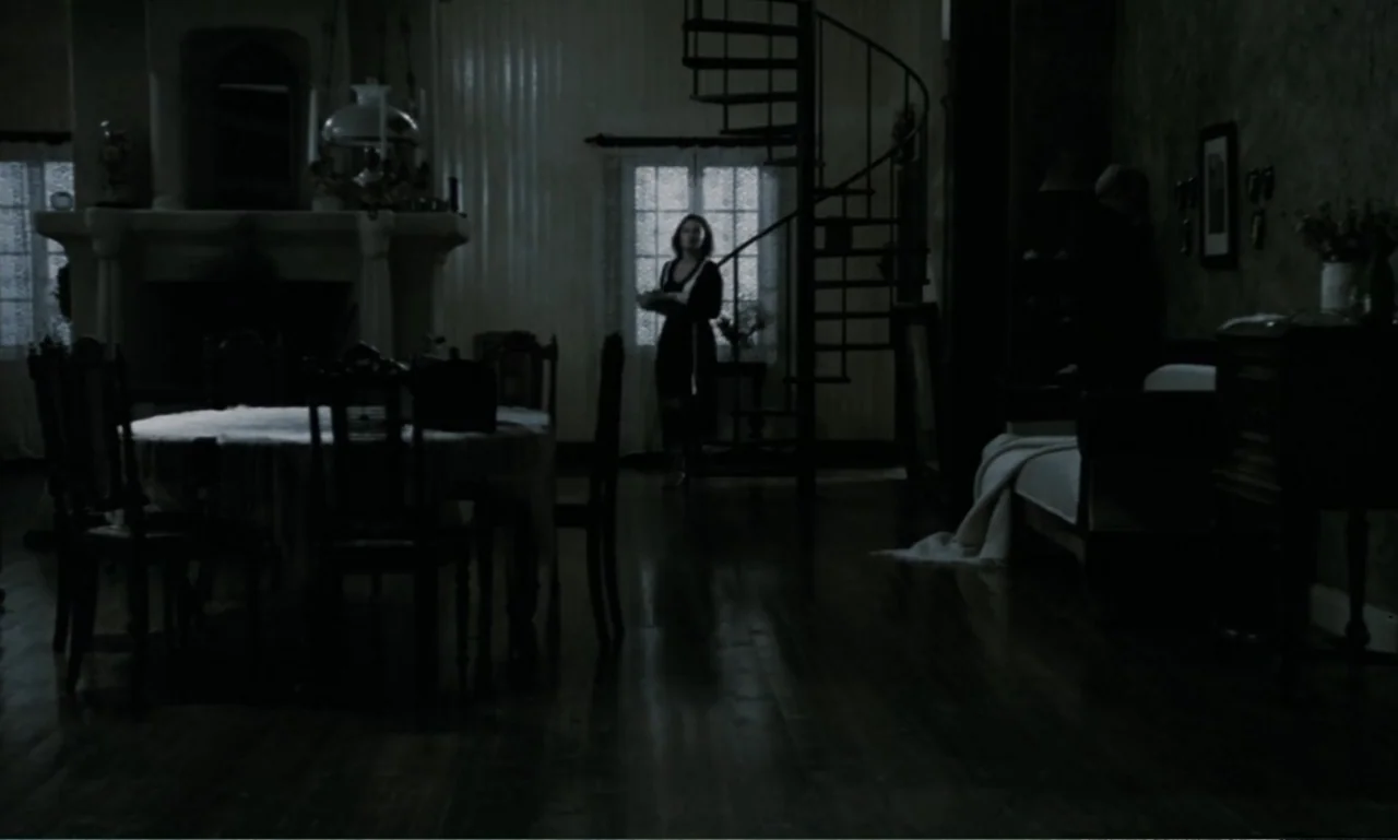 The Sacrifice (2015) movie still: wide — A dark, sparsely furnished room contains a dining set and a central spiral staircase. A woman wearing a…