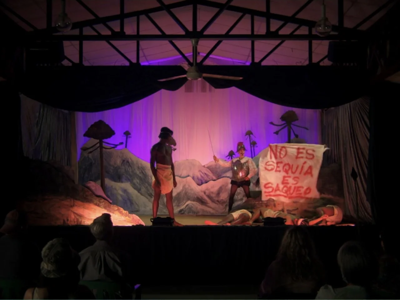 History and Geography (2024) movie still: wide — A theatrical production features actors on a painted landscape set with a protest sign. One…