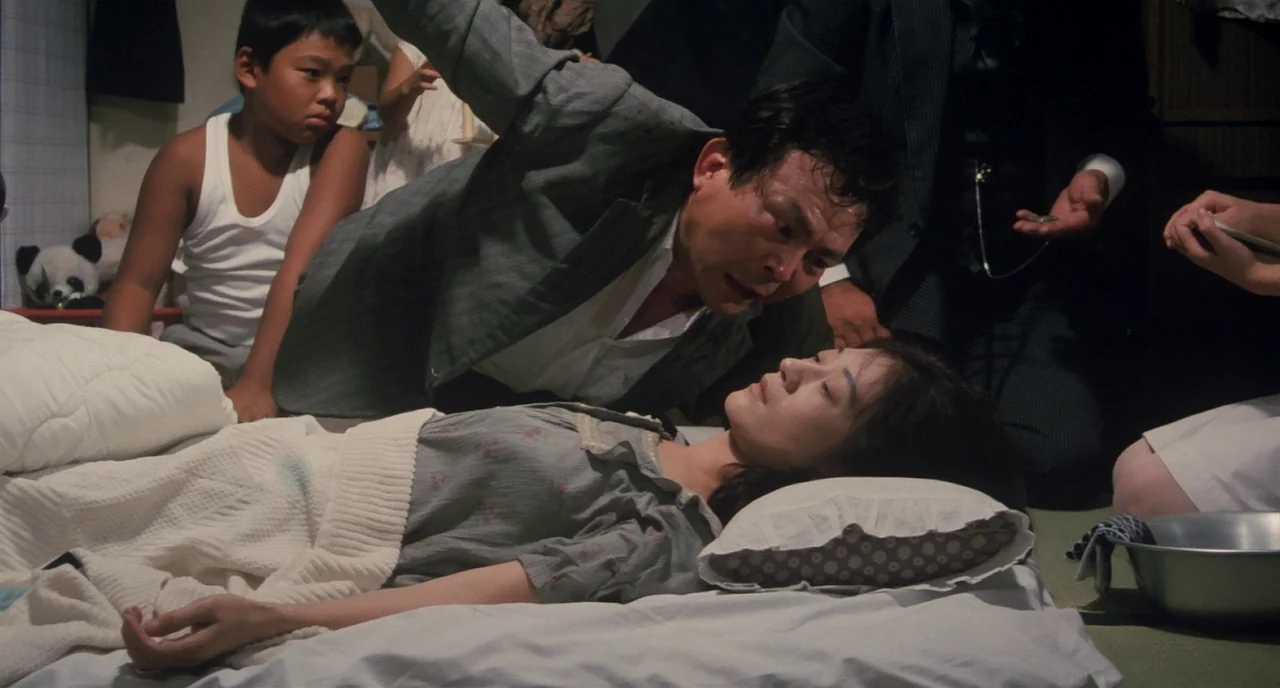 Tampopo (1985) movie still: medium — A Japanese interior with floor mats features a woman resting on a white futon. A man in a jacket leans over her…