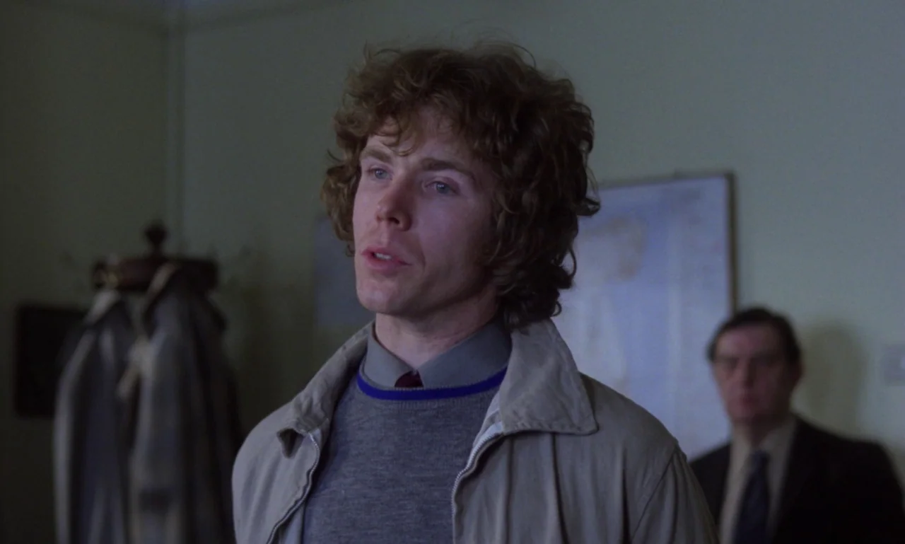 Scum (1979) movie still: medium — A spare, light-walled room features a rack of coats in the background corner. The main subject stands centered…
