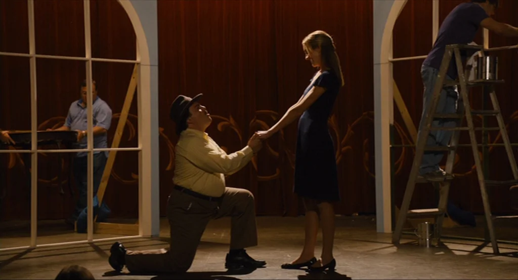 Bernie (2011) movie still: fullbody — A theatrical stage with red curtains in the background and white arched scenic frames. A man in a hat kneels…
