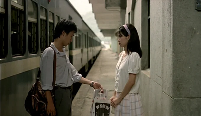 Dust In The Wind (1986) movie still: fullbody — A concrete train platform sits beside a long, stationary passenger train on a cloudy day. A man…