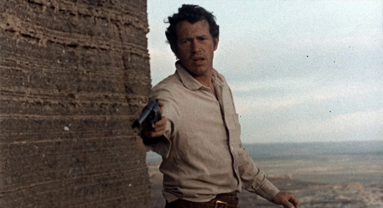The Shooting (1966) movie still: medium — A man stands on a desolate cliffside with layered rock formations behind him. He holds a revolver pointed…