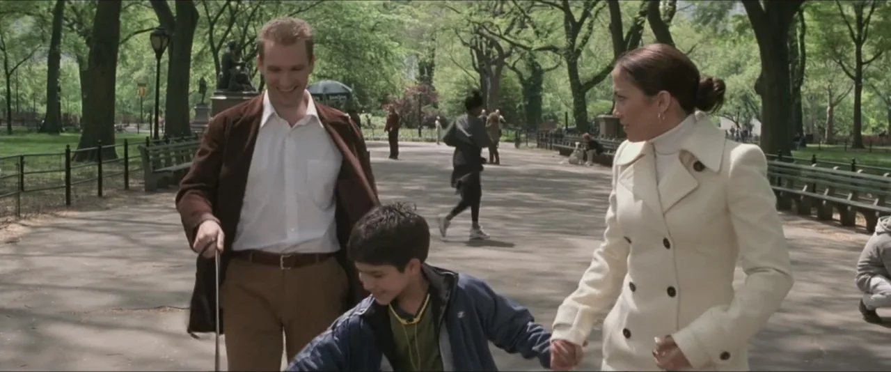 Maid in Manhattan (2002) movie still: fullbody — A paved path winds through a leafy public park with mature trees lining the route. A man and a…