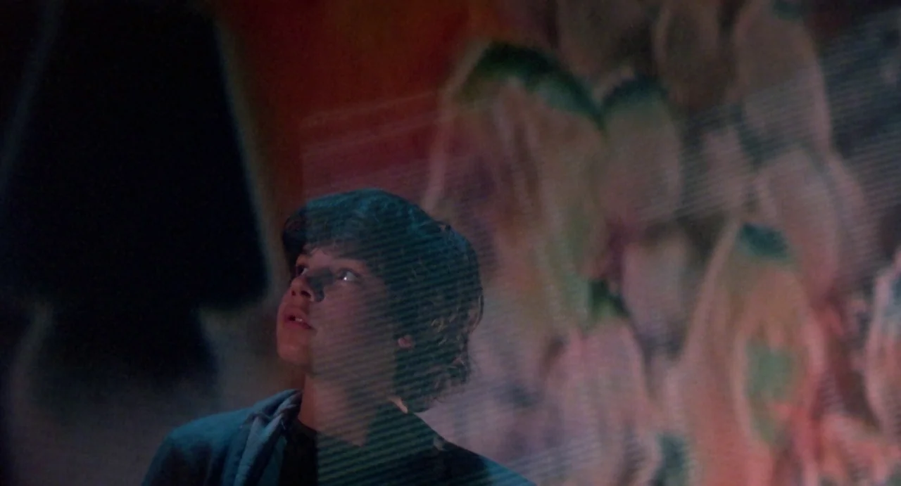 Explorers (1985) movie still: closeup — A teenage boy stands in a dark space, his face partially obscured by a projected image. A horizontal…