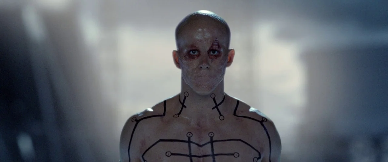 Deadpool 2 (2018) movie still: medium — A dimly lit industrial room contains a pale man with black ink markings. The man stands motionless with his…