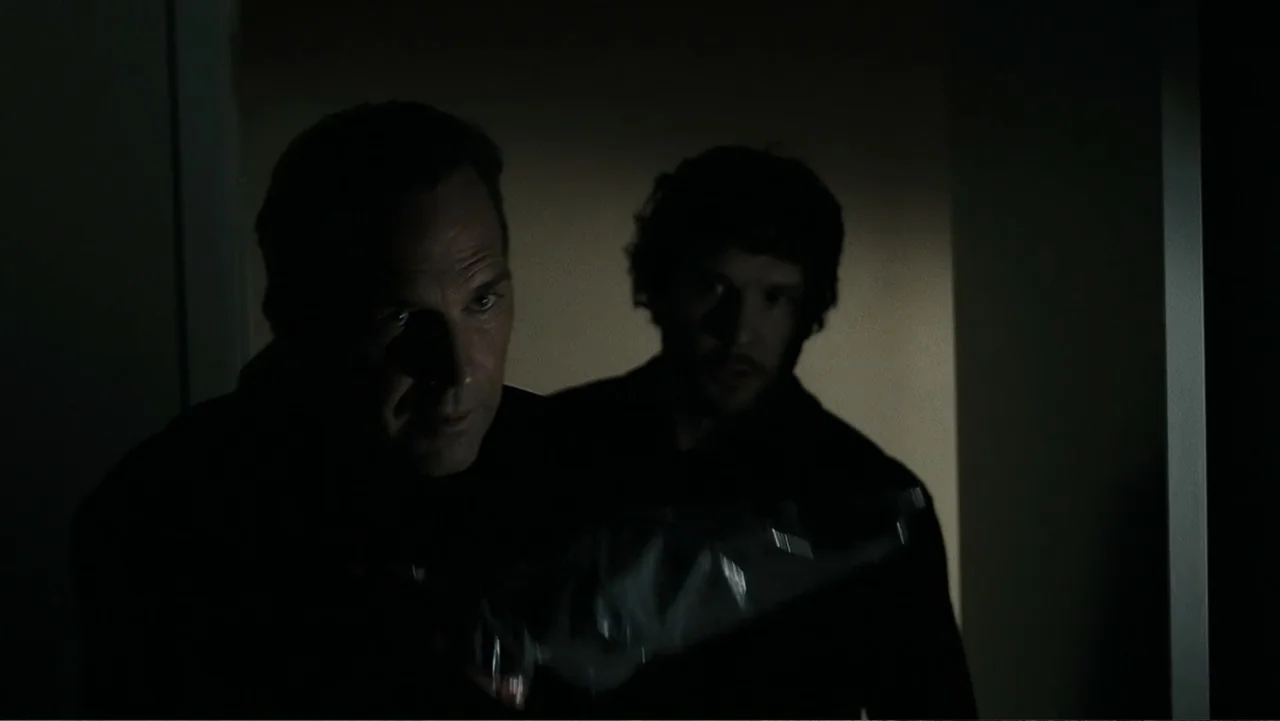 Mr. Brooks (2007) movie still: medium — A dark, dimly lit interior hallway with nondescript wall surfaces. Two men stand in the shadows and look…