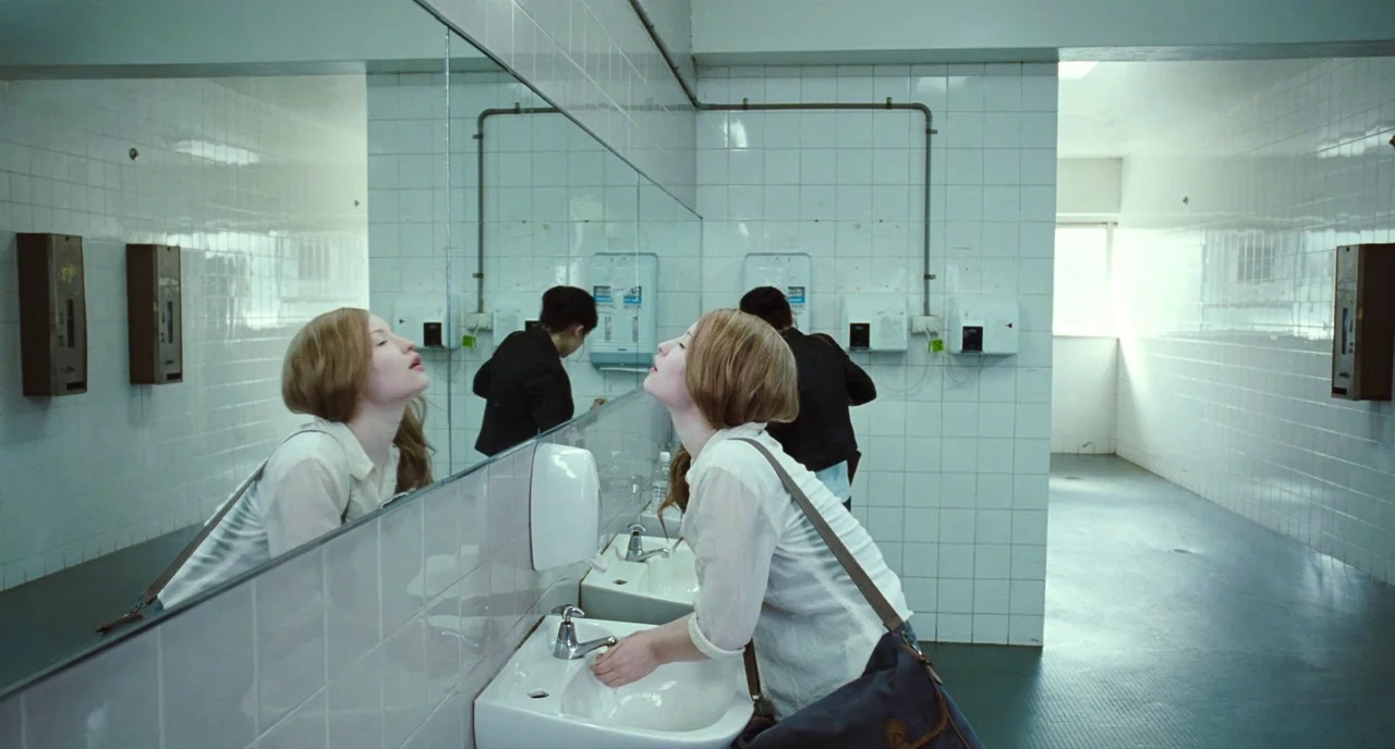 Sleeping Beauty (2011) movie still: medium — A sterile white tiled restroom features a long wall mirror and white porcelain basins. A young woman…