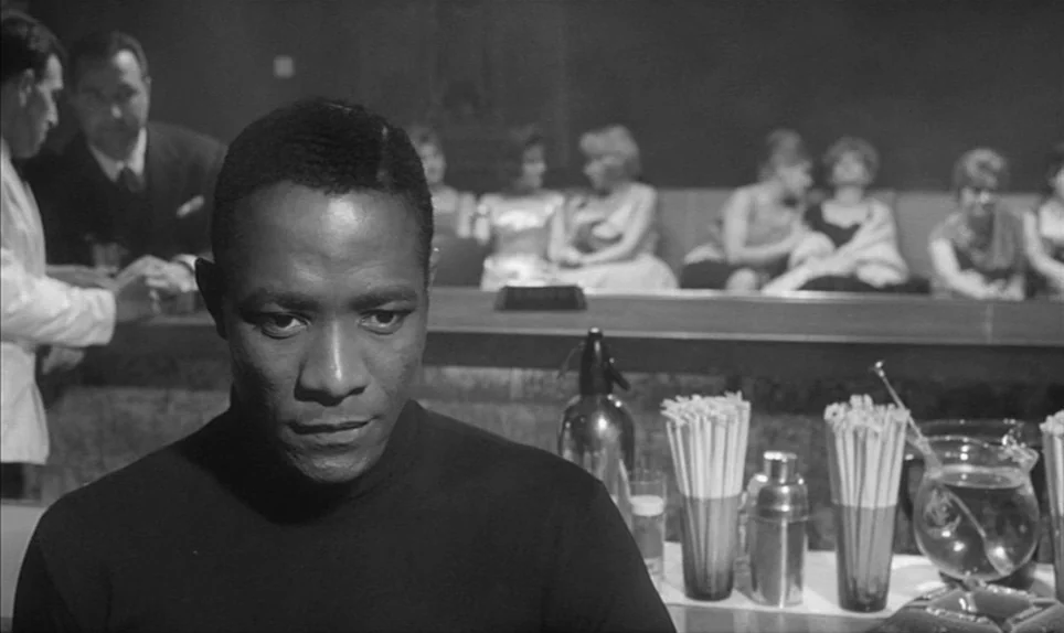 Eva (1962) movie still: medium — A dimly lit lounge with patrons seated on a booth in the background. The man sits at the bar counter with cocktail…