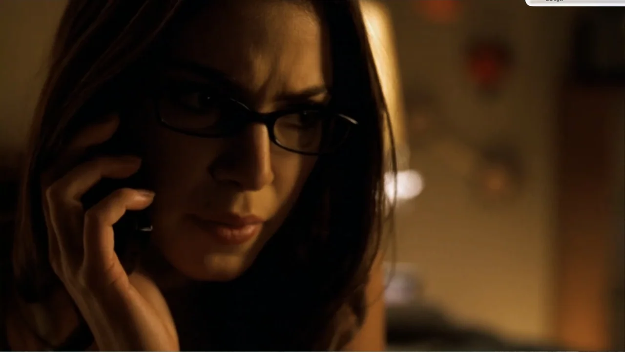 Chain Letter (2010) movie still: closeup — A dimly lit room with a soft out-of-focus background light in the dark. A woman with glasses speaks on a…