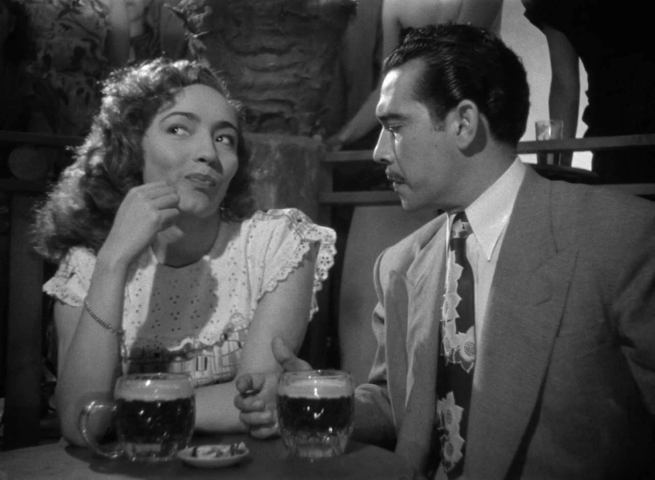 Victims of Sin (1951) movie still: medium — A crowded bar setting with a small round table in the foreground. A man and woman sit across from each…
