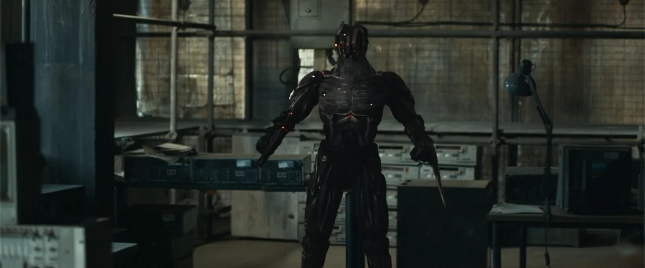 The Condemned 2 (2015) movie still: fullbody — An industrial workspace filled with metal shelving units and scattered electronic components. A dark…