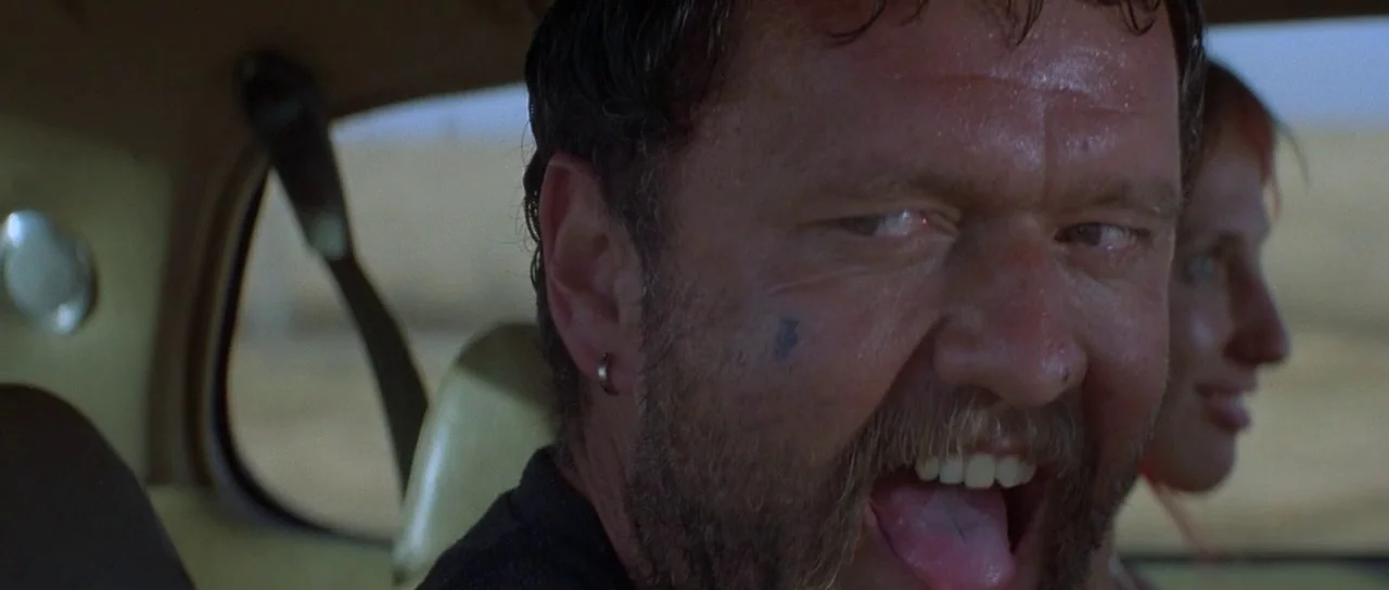 Mad Max (1979) movie still: closeup — The interior of a moving car during a bright day in an arid landscape. A man with a stubbly beard and a tattoo…