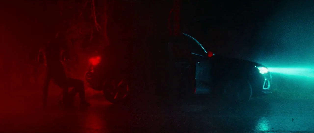 Inexorable (2021) movie still: wide — A dark, wet asphalt road surface at night with thick atmospheric haze present. A silhouetted person sits on…