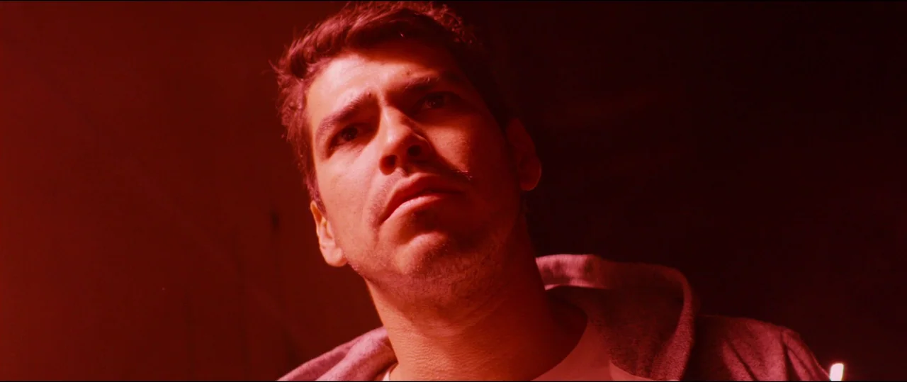 El Chicano (2018) movie still: closeup — A man stands in an dark, empty space illuminated by intense red light. He gazes upward at an unseen object.