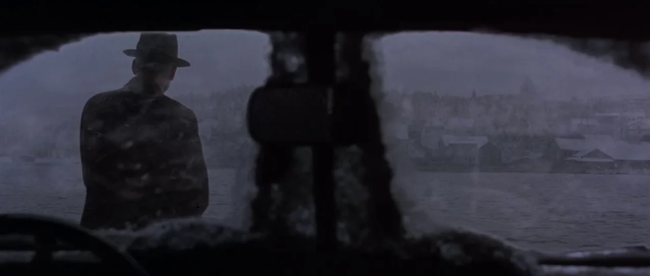 Snow Falling on Cedars (1999) movie still: wide — A man stands on a snowy dock overlooking a cold, grey body of water. Houses line the distant,…