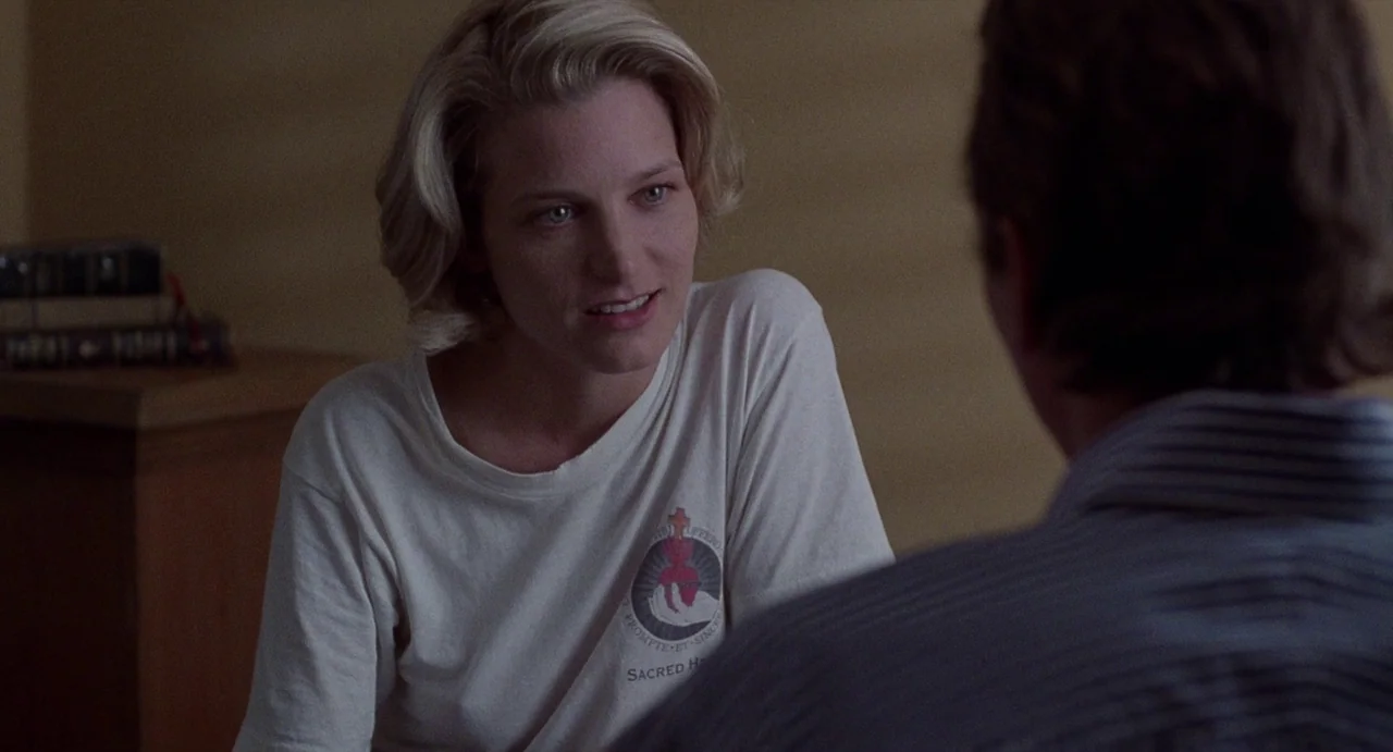 Touch (1997) movie still: medium — A woman with short blonde hair sits inside a dimly lit domestic space. She faces a man seen from behind who wears…