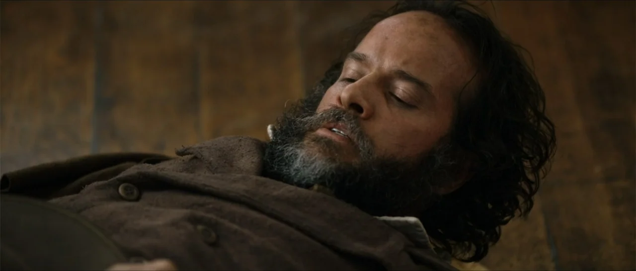 Out of Liberty (2019) movie still: closeup — A man lies on a rustic wooden plank floor in a dark interior space. The man wears a heavy brown coat…