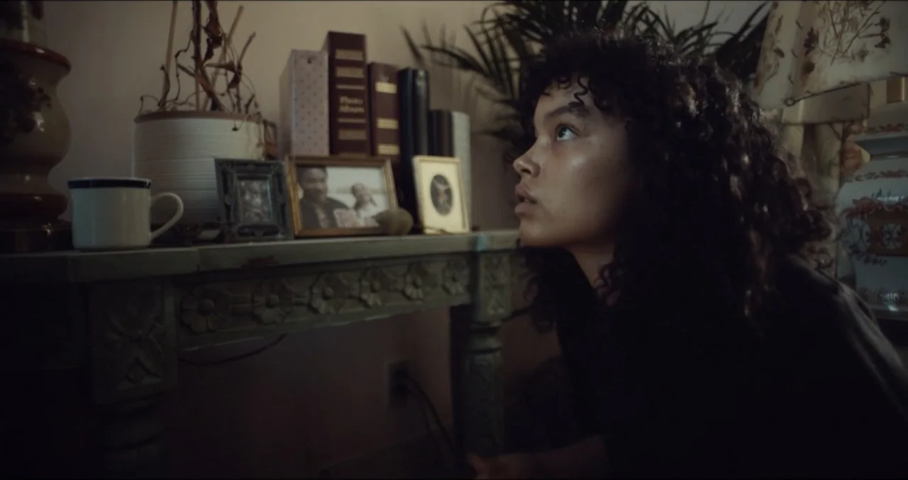 Eye for an Eye (2025) movie still: medium — A living room interior features a carved wooden table topped with books and photographs. A young woman…