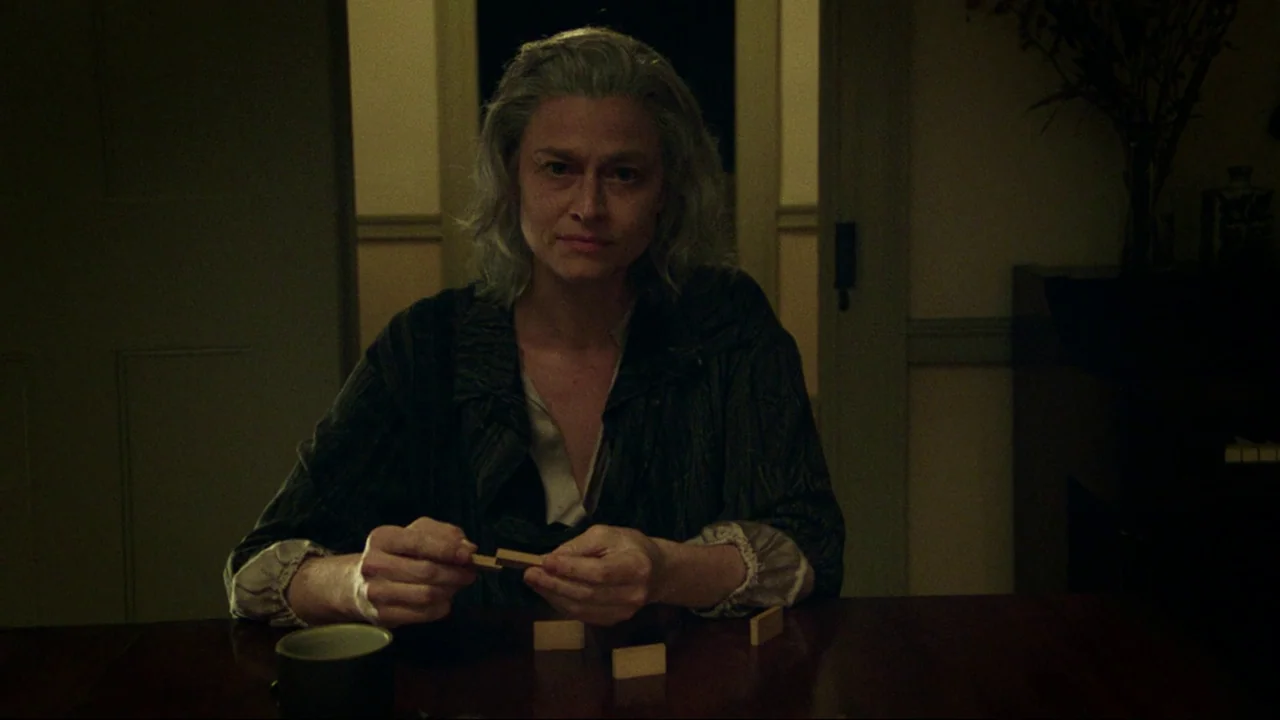 After Yang (2021) movie still: medium — A dimly lit room with muted earth tones and dark doorways in the background. A woman sits at a table and…