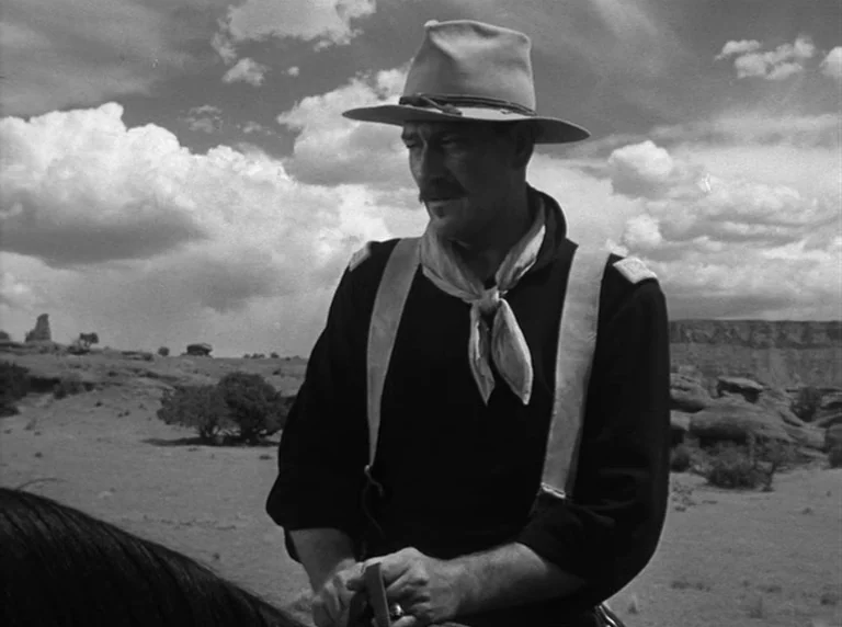 Rio Grande (1950) movie still: medium — A dry desert landscape with low scrub brush and distant mesas under a clouded sky. A man in a brimmed hat…