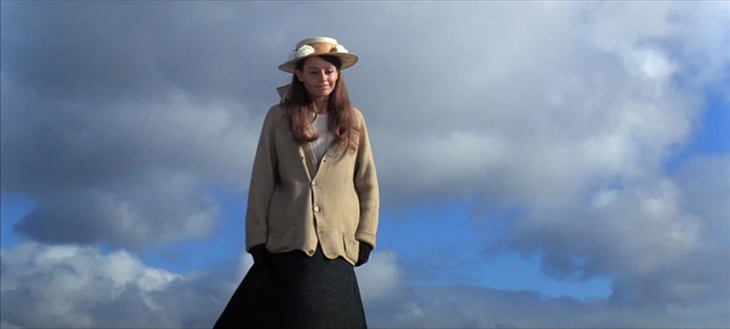 Ryan’s Daughter (1970) movie still: fullbody — A woman stands against a backdrop of clouds and bright blue sky. She wears a straw hat and a knit…