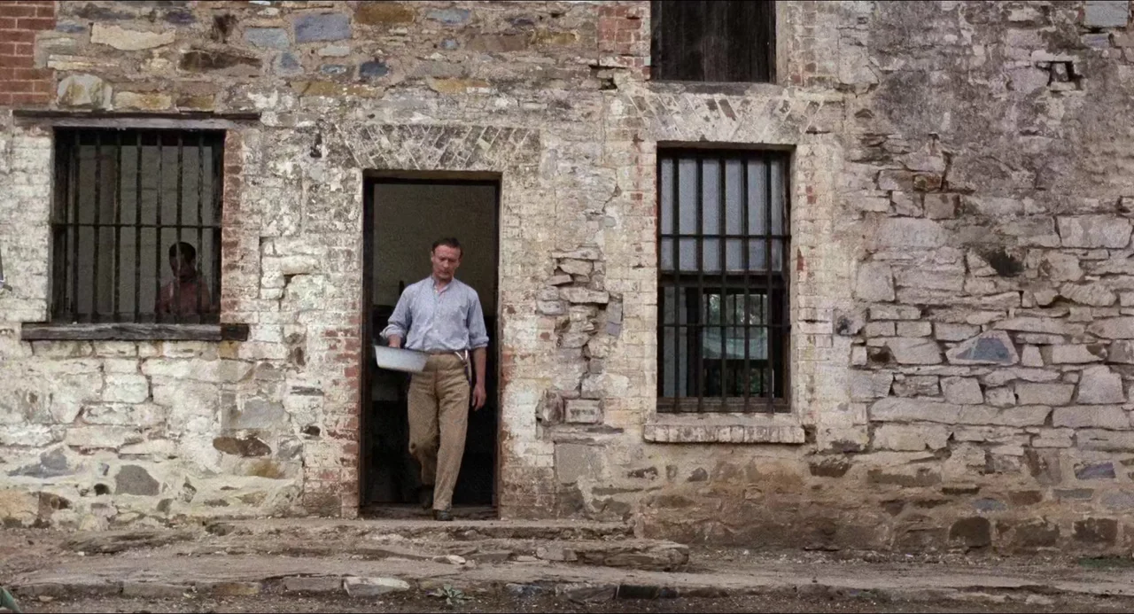 Breaker Morant (1980) movie still: fullbody — A weathered stone wall features barred windows and a central dark doorway. A man walks through the…