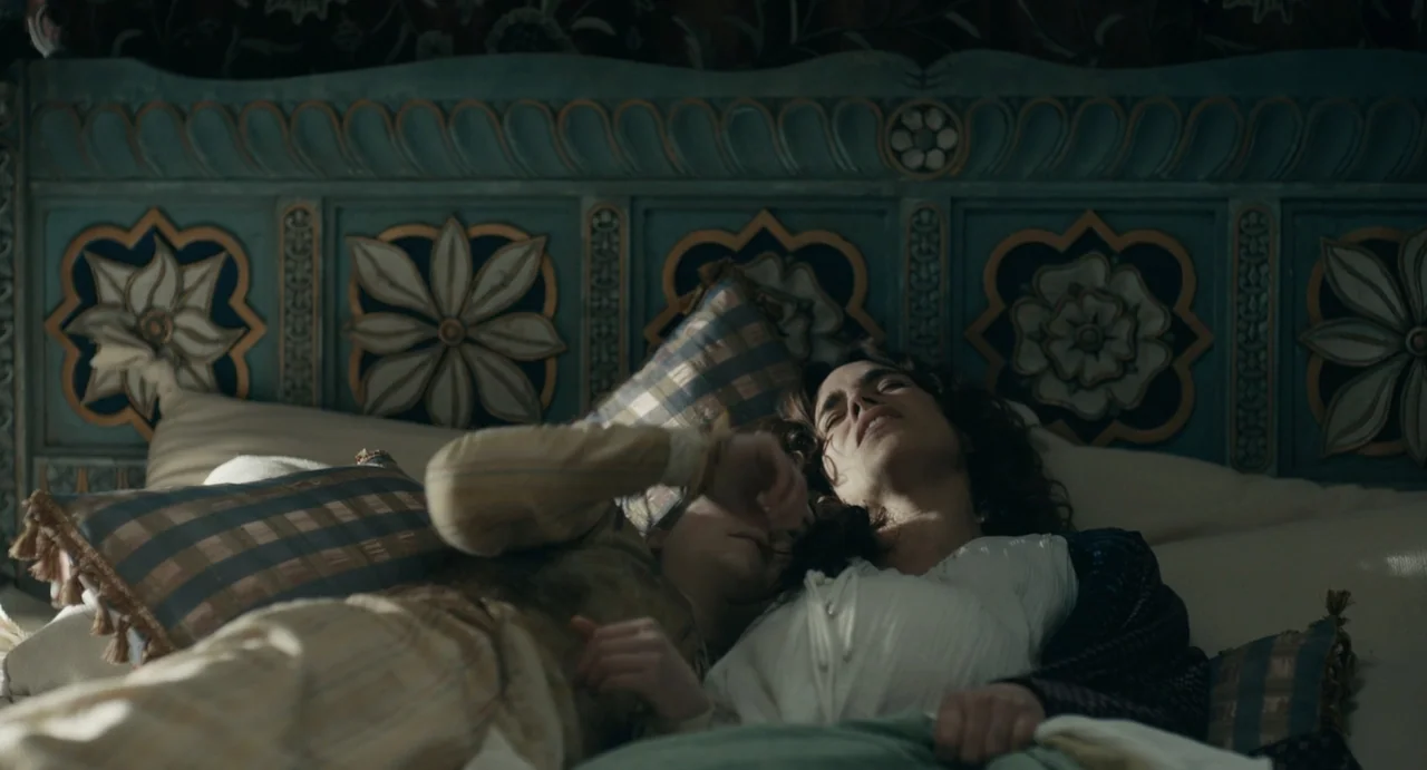 Catherine Called Birdy (2022) movie still: medium — Two women recline together on a bed with an ornate blue wooden headboard. The women lie close…