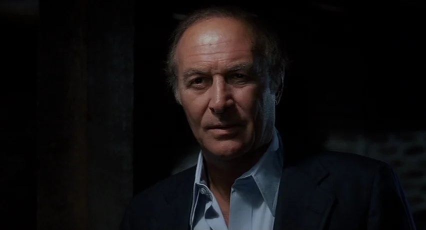 Psycho 2 (1983) movie still: closeup — A dimly lit room with a dark wooden support beam visible in the background. A mature man looks directly at…