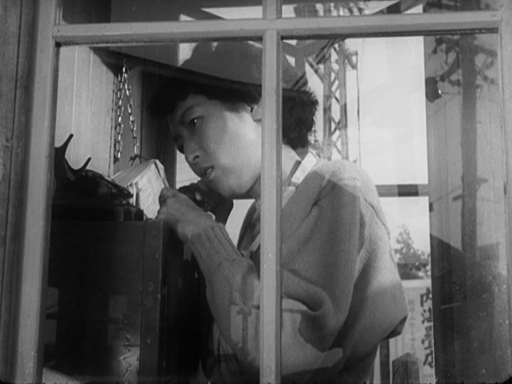 Ikiru (1952) movie still: medium — A woman stands inside a small glass phone booth during the day. She speaks into a vintage telephone receiver…