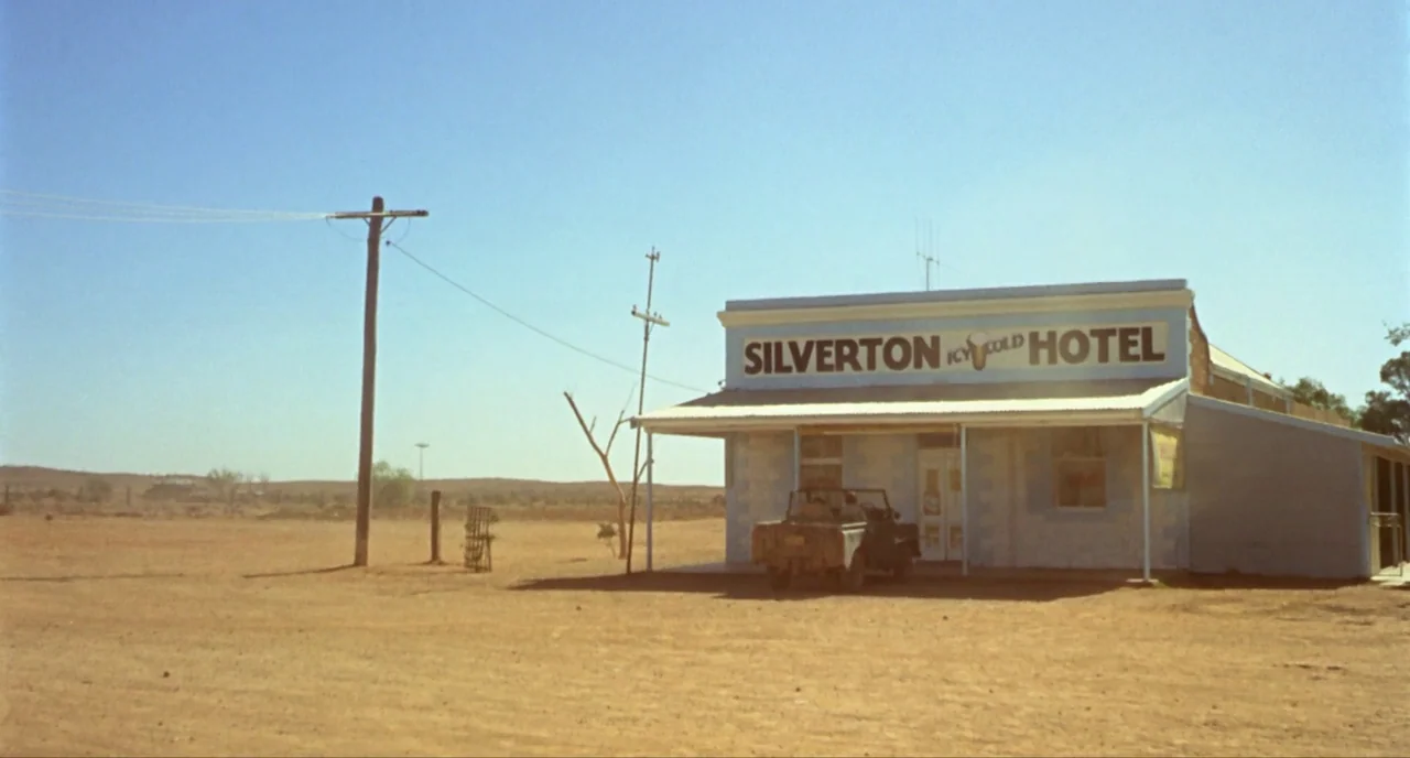 Wake In Fright (1971) movie still: establishing — A single-story building sits in a dry, barren desert landscape under a clear sky. A vintage…