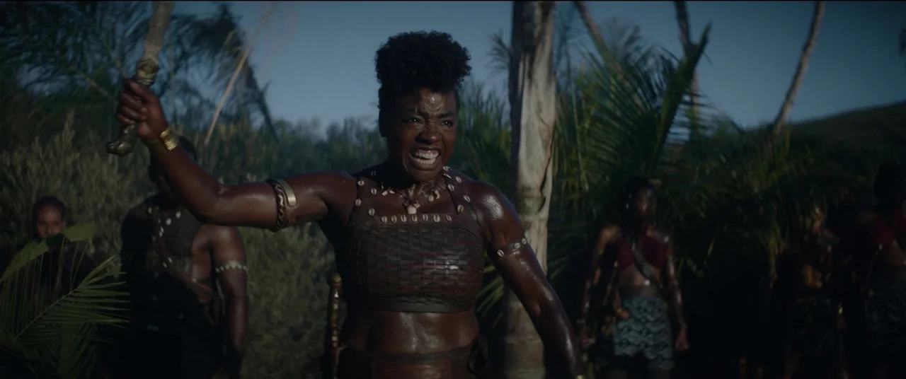 The Woman King (2022) movie still: medium — A group of warriors stands within a dense thicket of palm trees and tropical brush. A woman in dark…