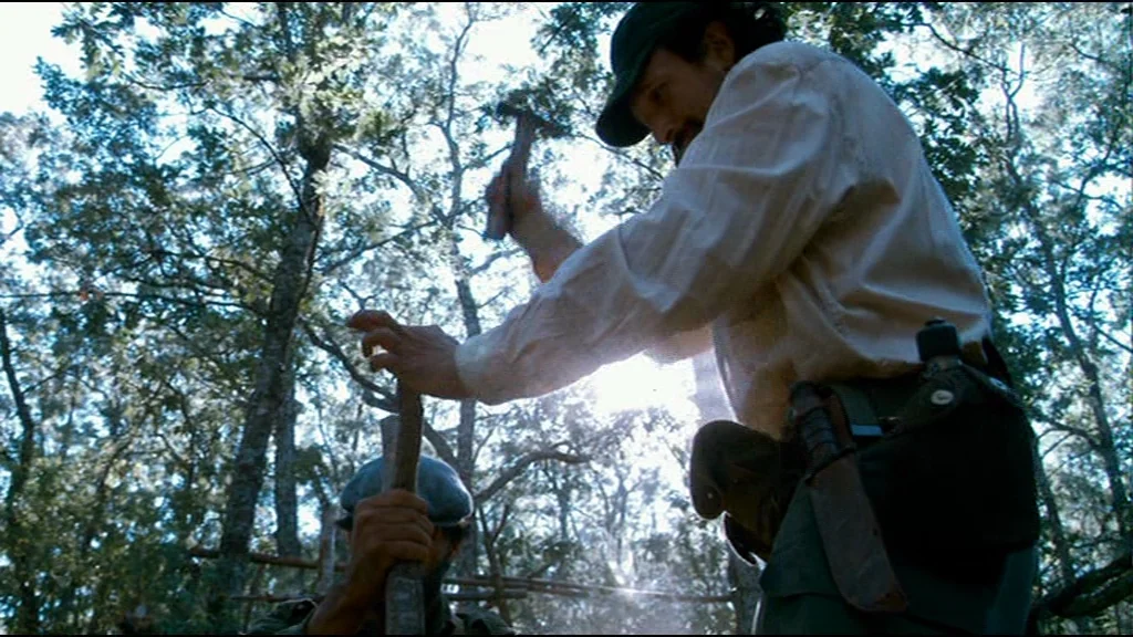 Che Part 2 (Guerrilla) (2008) movie still: medium — A sunlit forest clearing with dense trees and a wooden construction project in progress. A man…