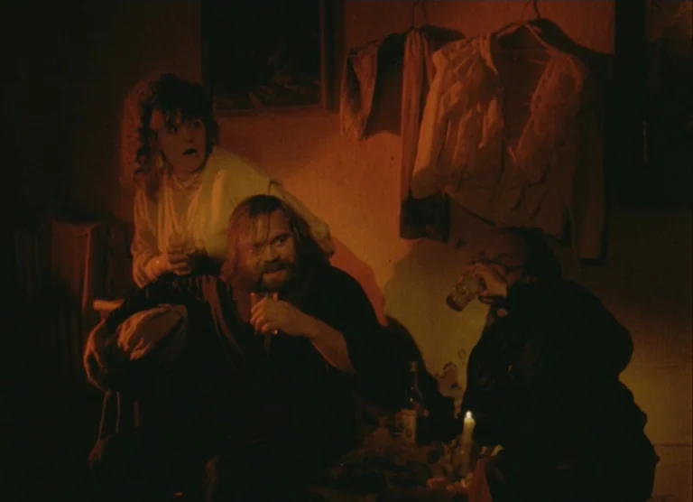 Russian Symphony (1994) movie still: medium — A dimly lit room features three people huddled together with hanging garments on the wall. The…