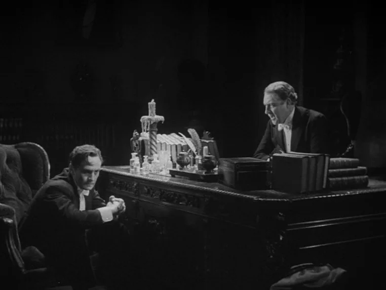 Dr. Jekyll and Mr. Hyde (1931) movie still: medium — A dark, wood-paneled room is dominated by a heavy, ornate desk. Two men in formal evening wear…