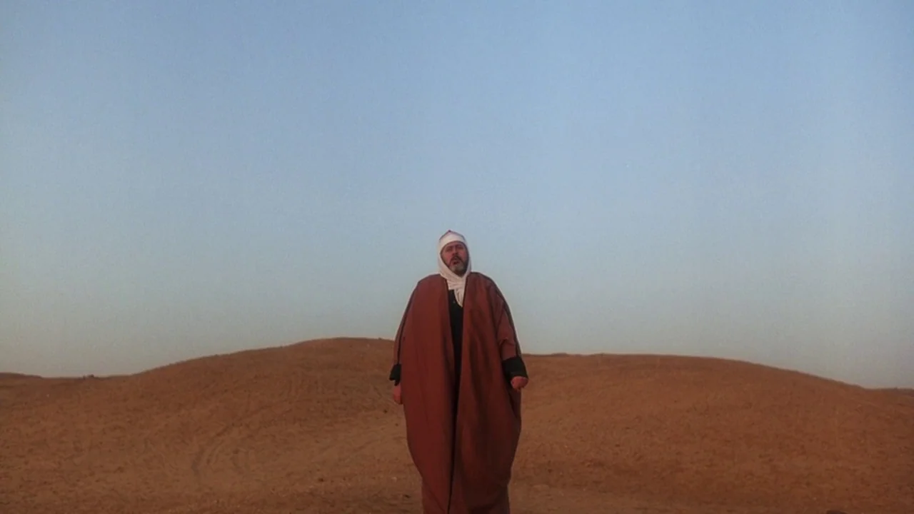 Malcolm X (1992) movie still: fullbody — An arid, rolling sandy landscape extends toward a clear horizon under a blue sky. A man wearing a headscarf…