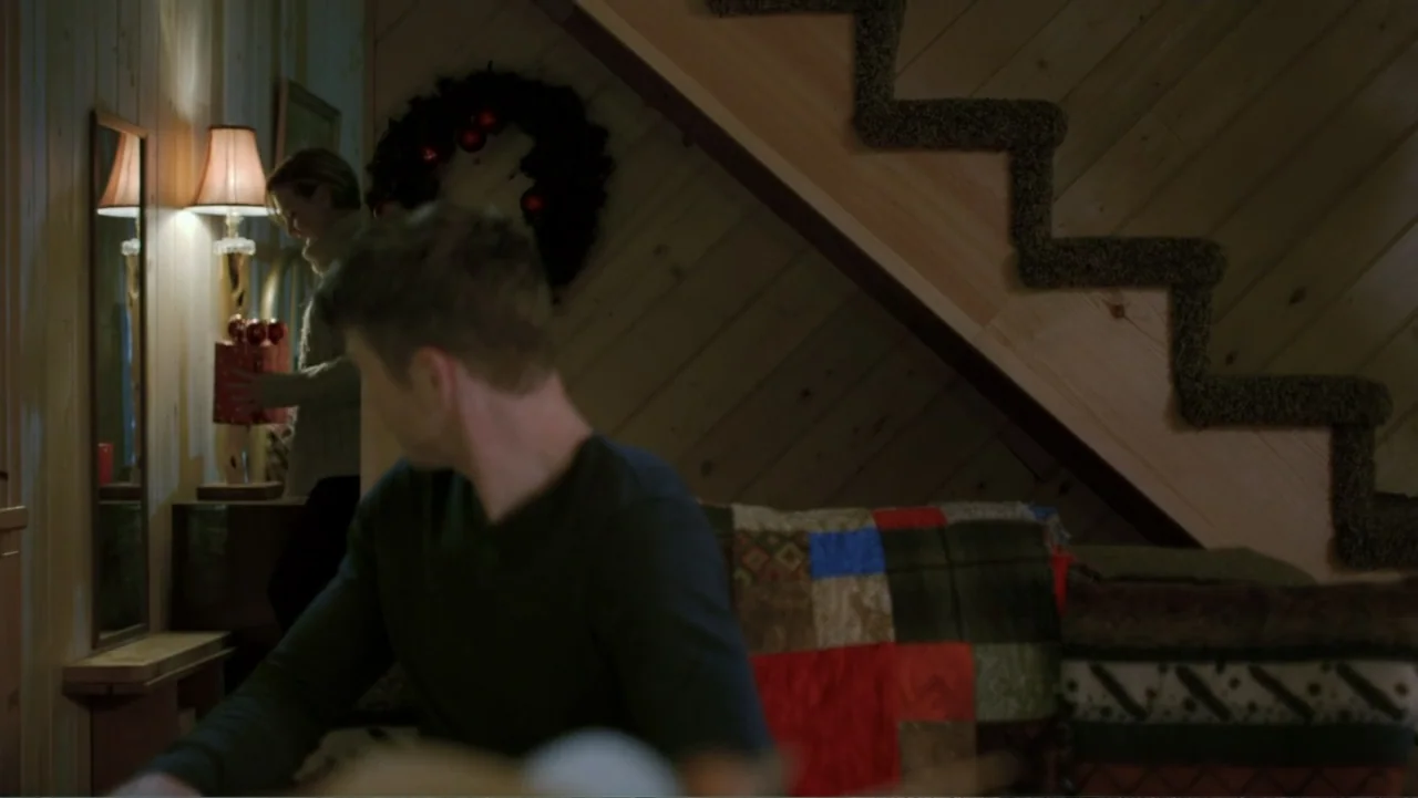 Alex & Emma (2003) movie still: medium — A living room with pine wood paneled walls and a carpeted staircase. A woman stands holding a gift near a…