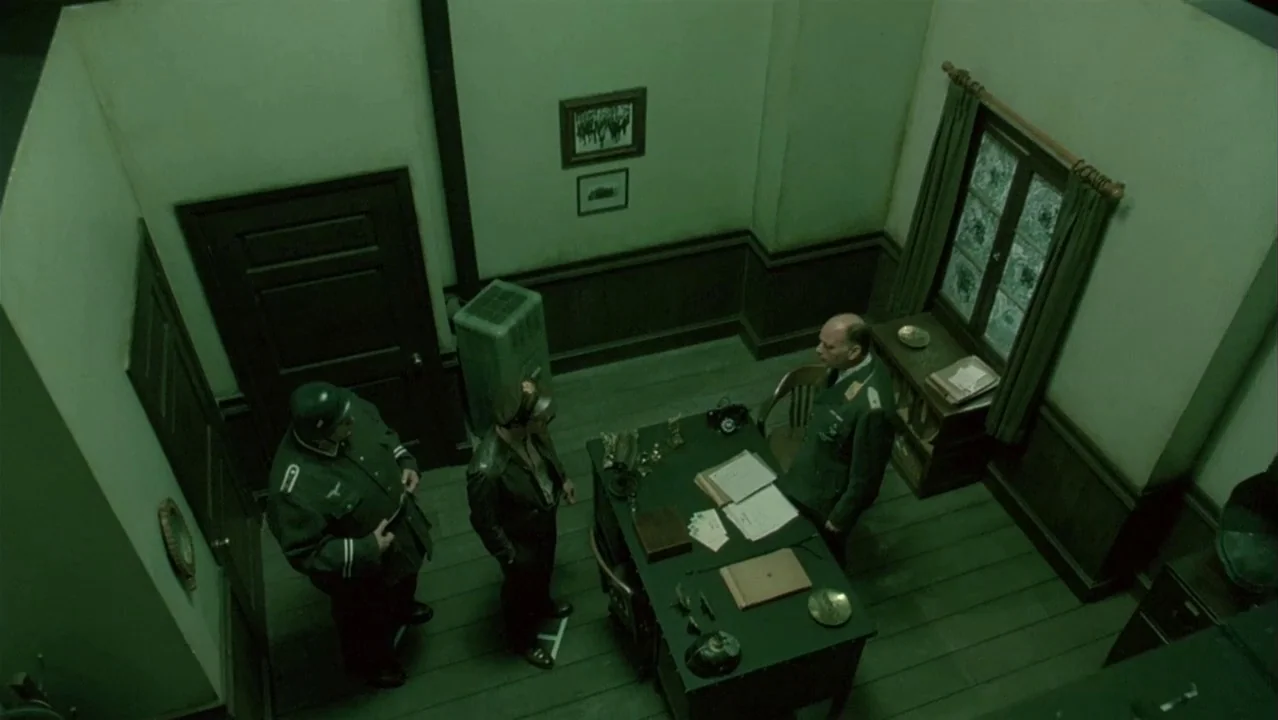 Auto Focus movie still: wide — A austere room featuring a desk with papers and a small window with curtains. Three men in military uniforms stand…