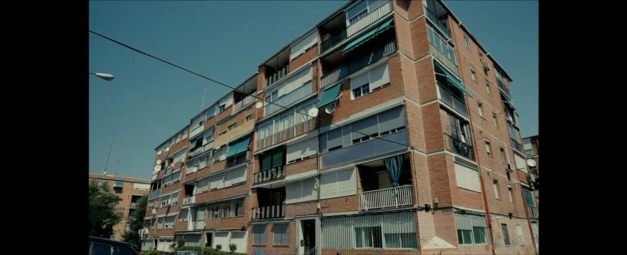 Faraday (2013) movie still: establishing — A multi-story red brick apartment building with multiple balconies and green fabric awnings. The…