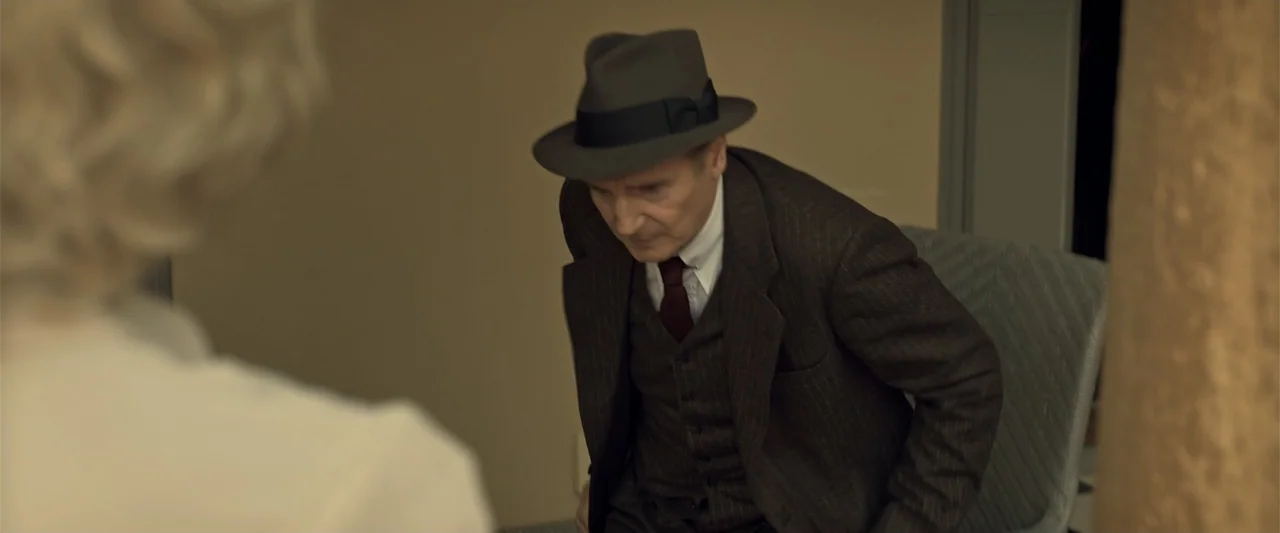 Marlowe (2023) movie still: medium — A dimly lit interior space with plain beige walls and a grey chair. An older man in a fedora and pinstripe suit…