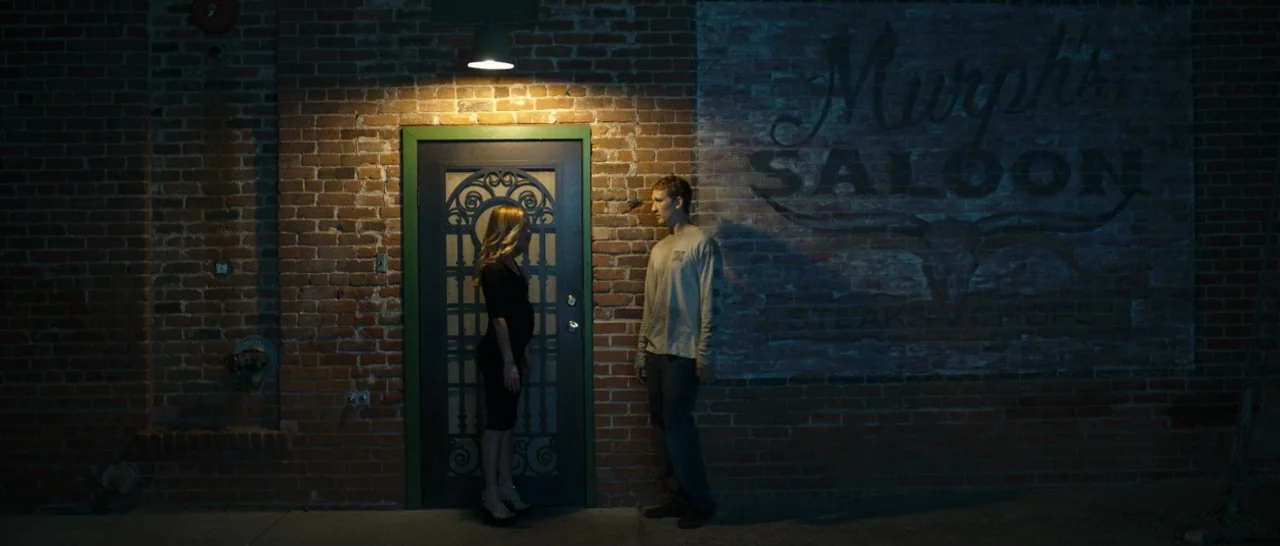 Only The Brave (2017) movie still: wide — A weathered brick building at night features a painted faded logo for a saloon. A man and a pregnant woman…