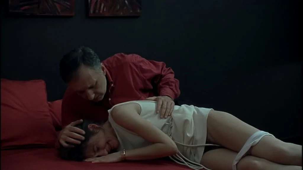Romance (1999) movie still: medium — A dark, minimally furnished room features a bright red bed frame and bedding. A man leans over a woman lying on…