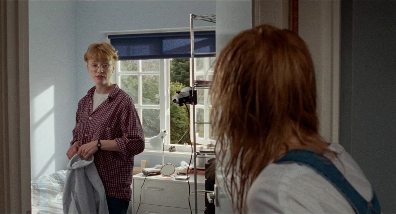 Life Is Sweet (1990) movie still: medium — A light blue bedroom with a window and a small metal shelf unit. A person with ginger hair stands in the…