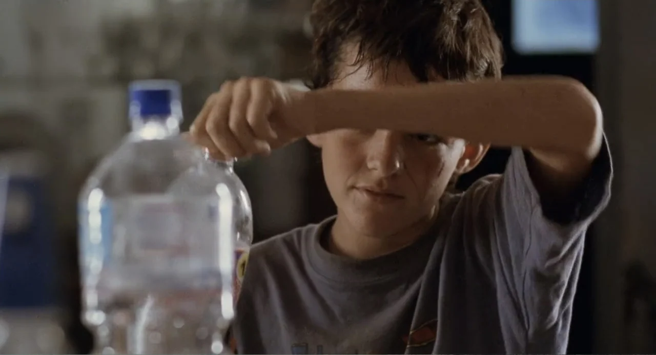 La Ciénaga (2001) movie still: closeup — A dimly lit indoor space with blurred background details. A young boy in a grey t-shirt opens a plastic…