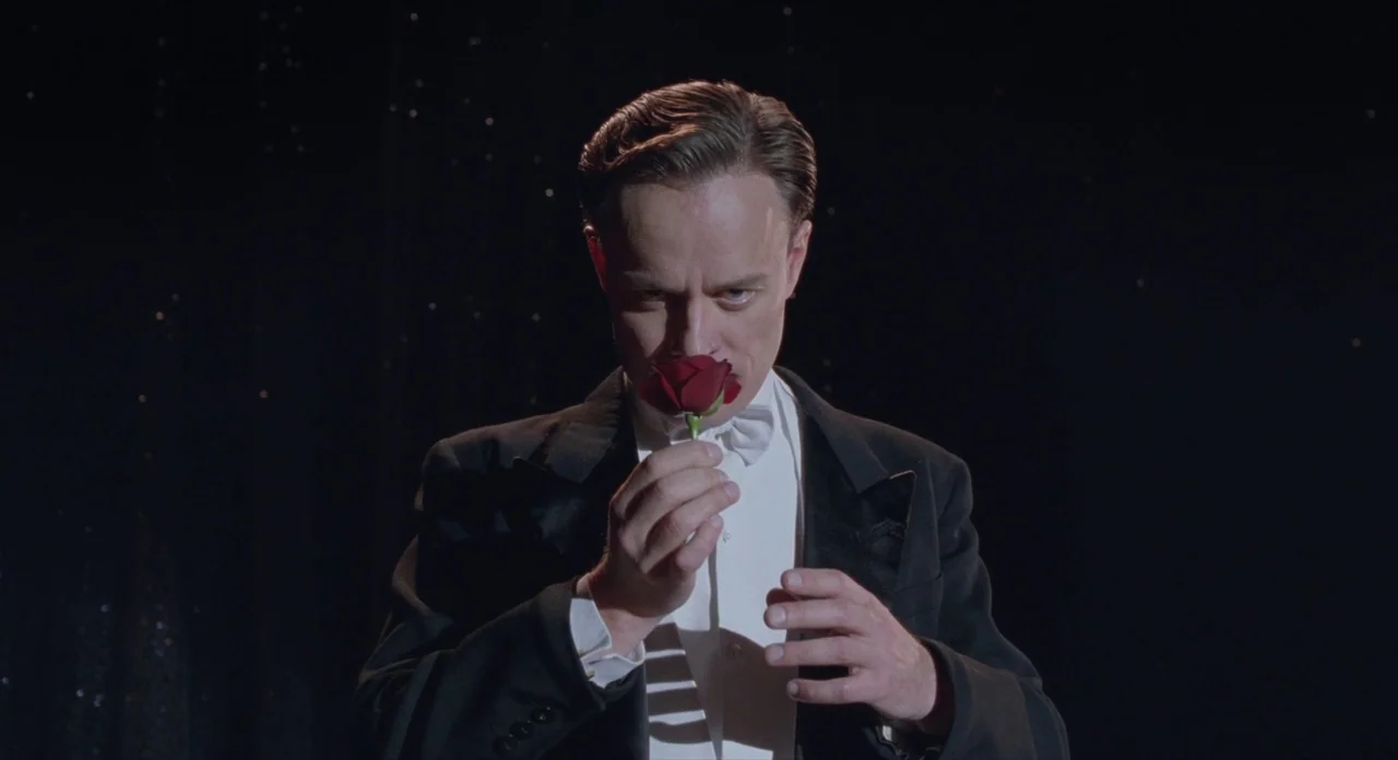 Populaire (2012) movie still: medium — A man in formal evening wear stands against a dark, featureless background. He holds a single red rose up to…
